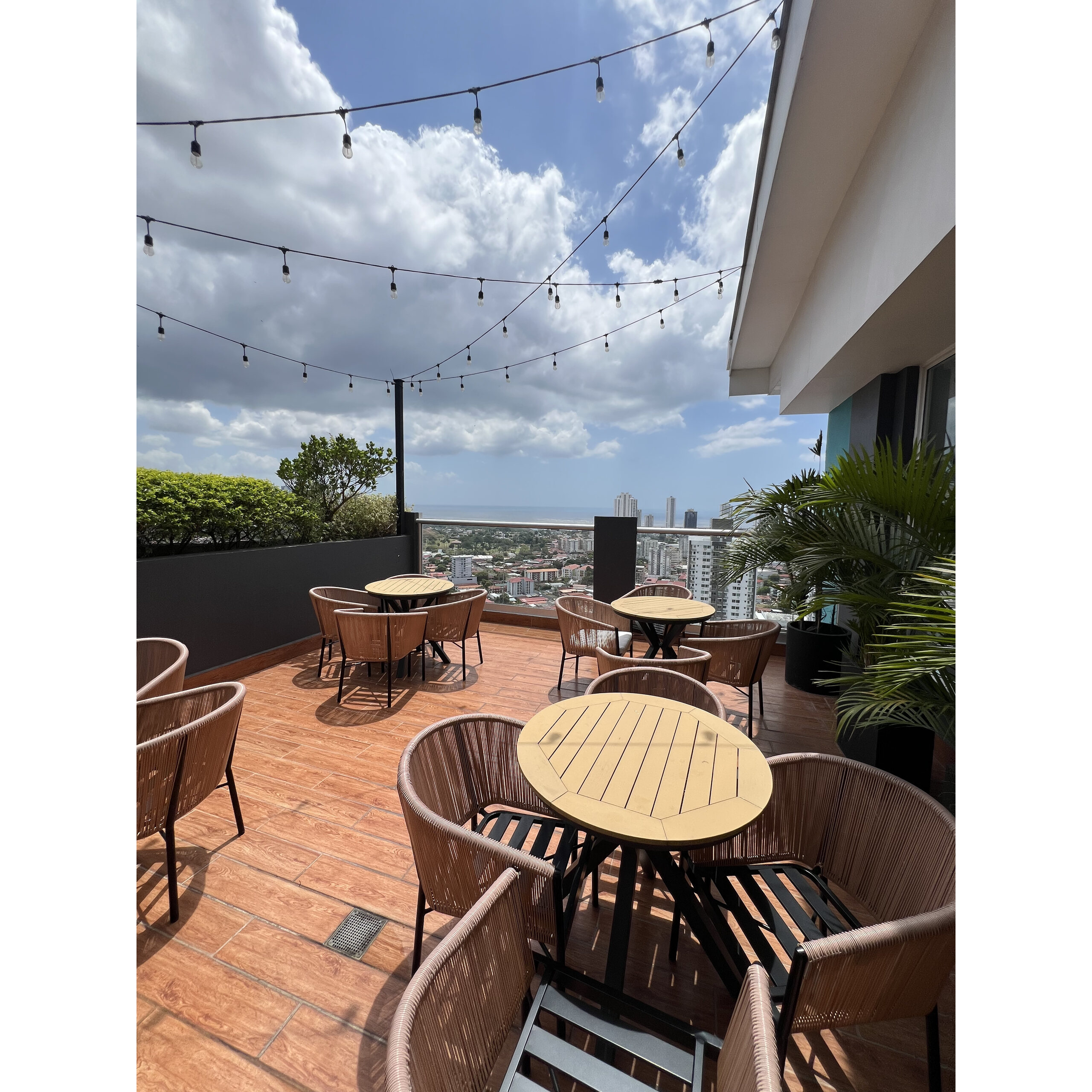 Terraza exterior con mesas y sillas, luces colgantes y vista urbana al fondo.
