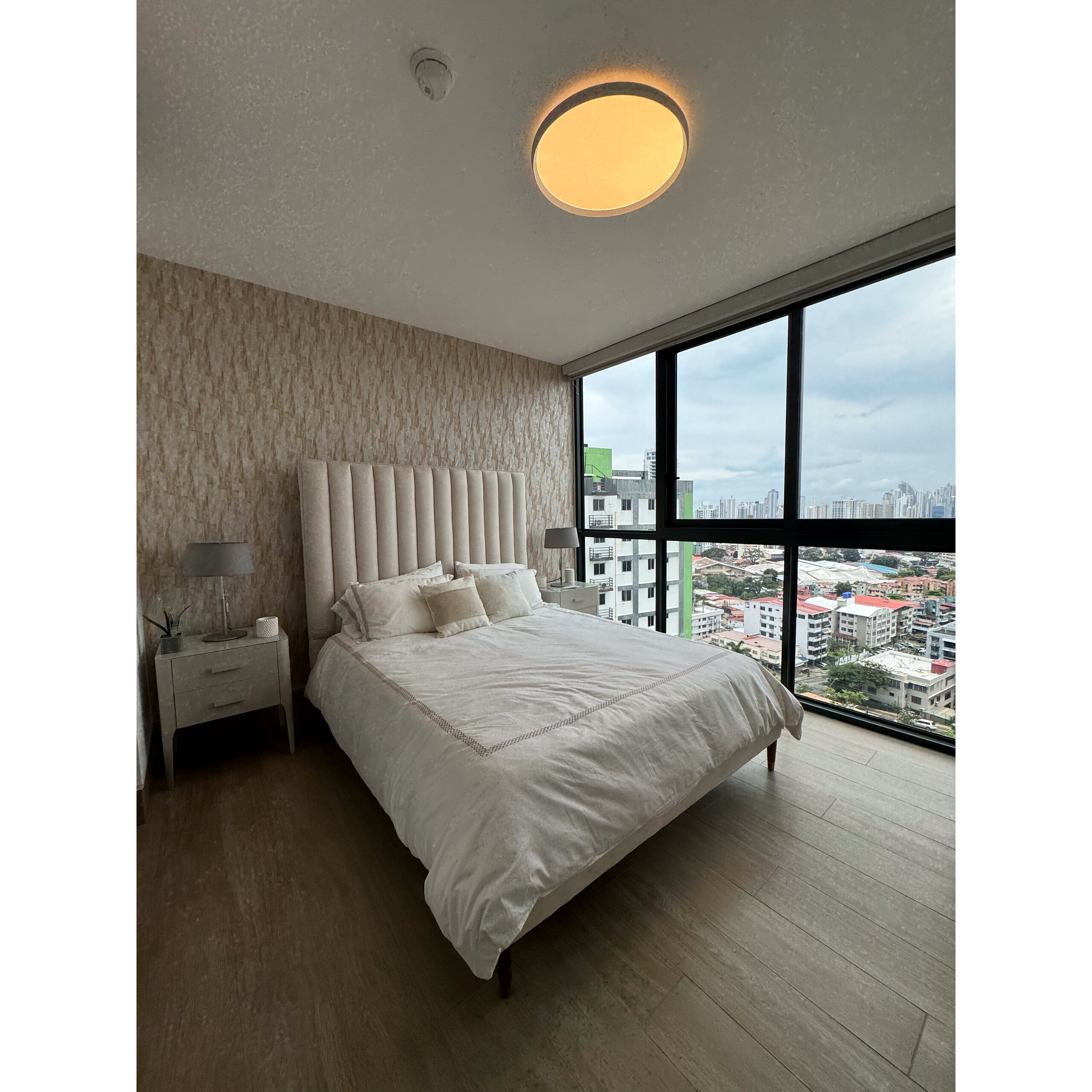 Recámara principal con cama, buró y ventanales con vista urbana.