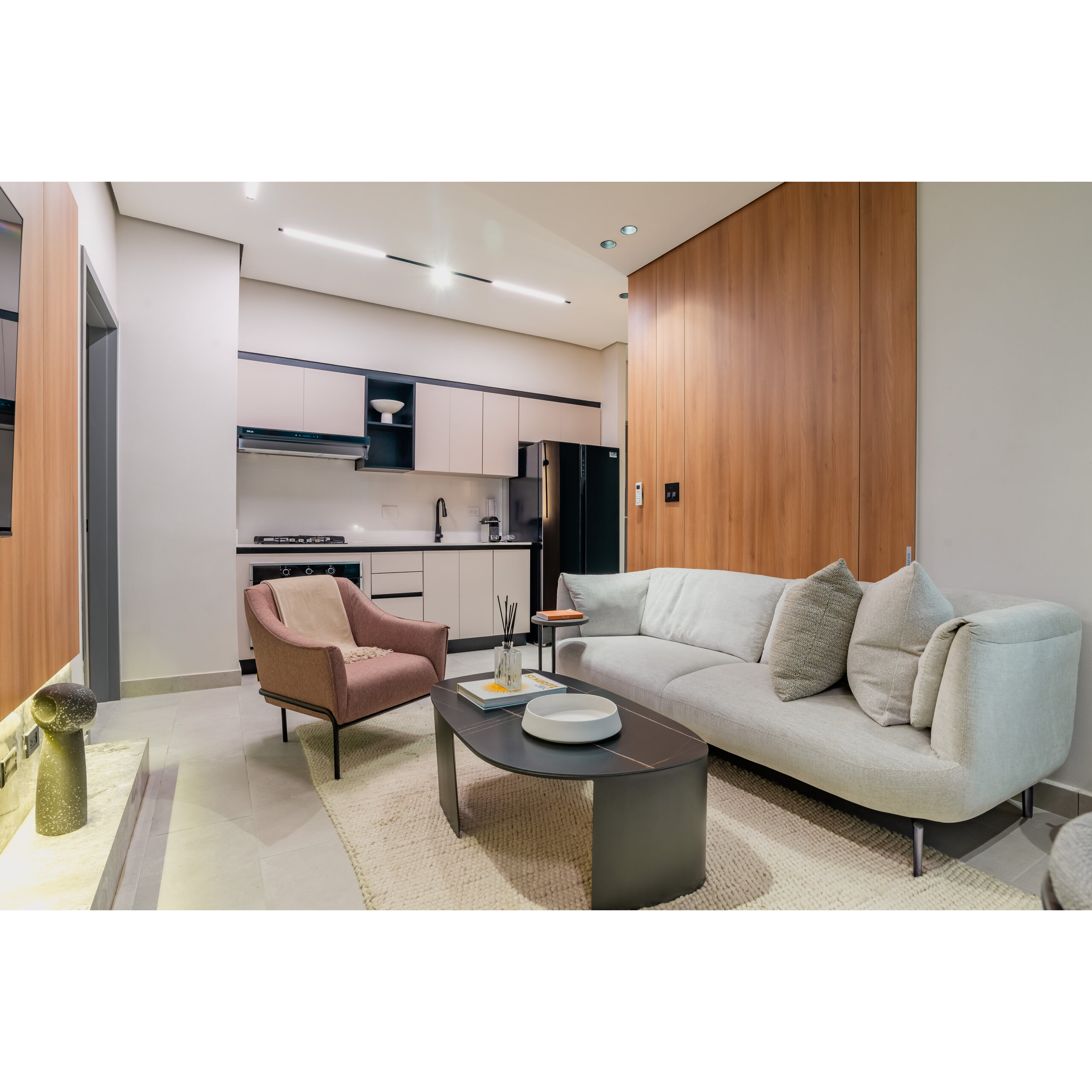 Sala con cocina abierta, sofá gris, sillón rosa y mesa de centro.