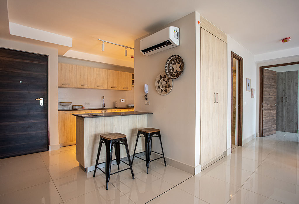 Área social con cocina abierta, barra y dos bancos, junto a un pasillo.