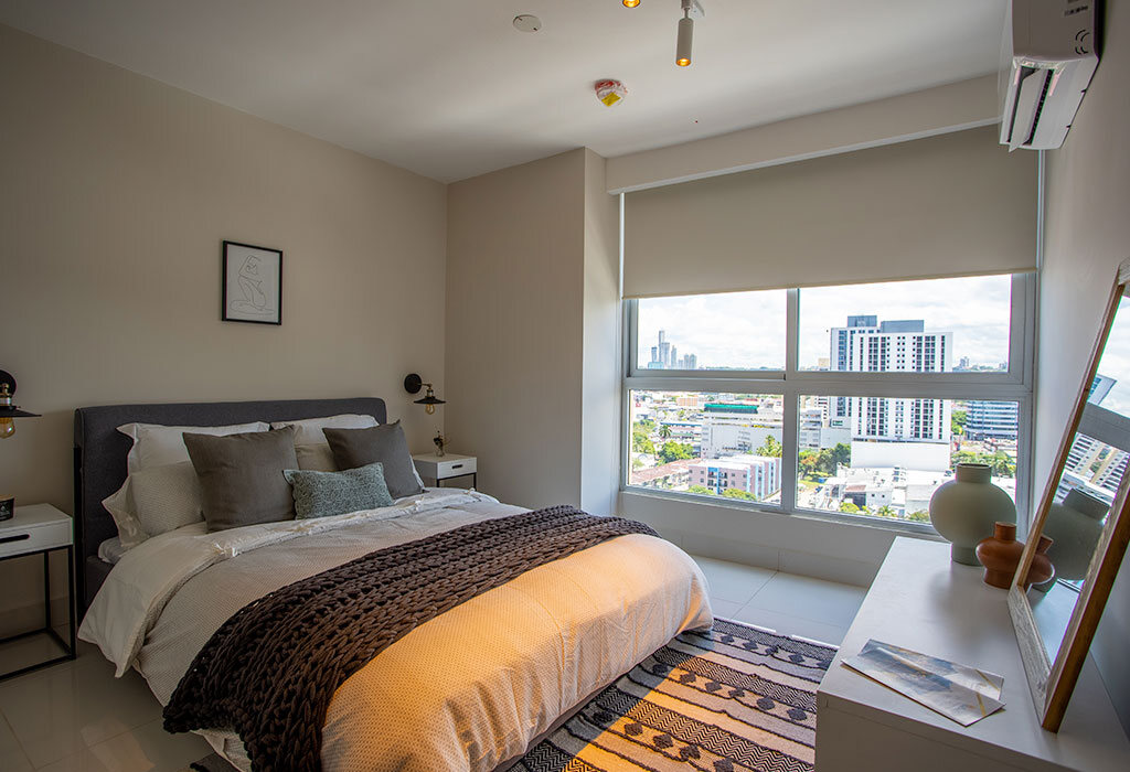 Recámara luminosa con cama, mesa de noche y gran ventana con vista urbana.