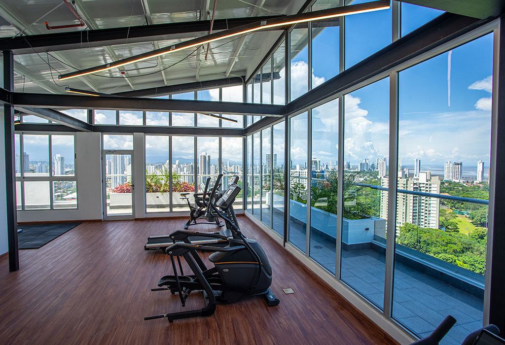 Área social con piso de madera, máquinas de ejercicio y ventanales con vista urbana.