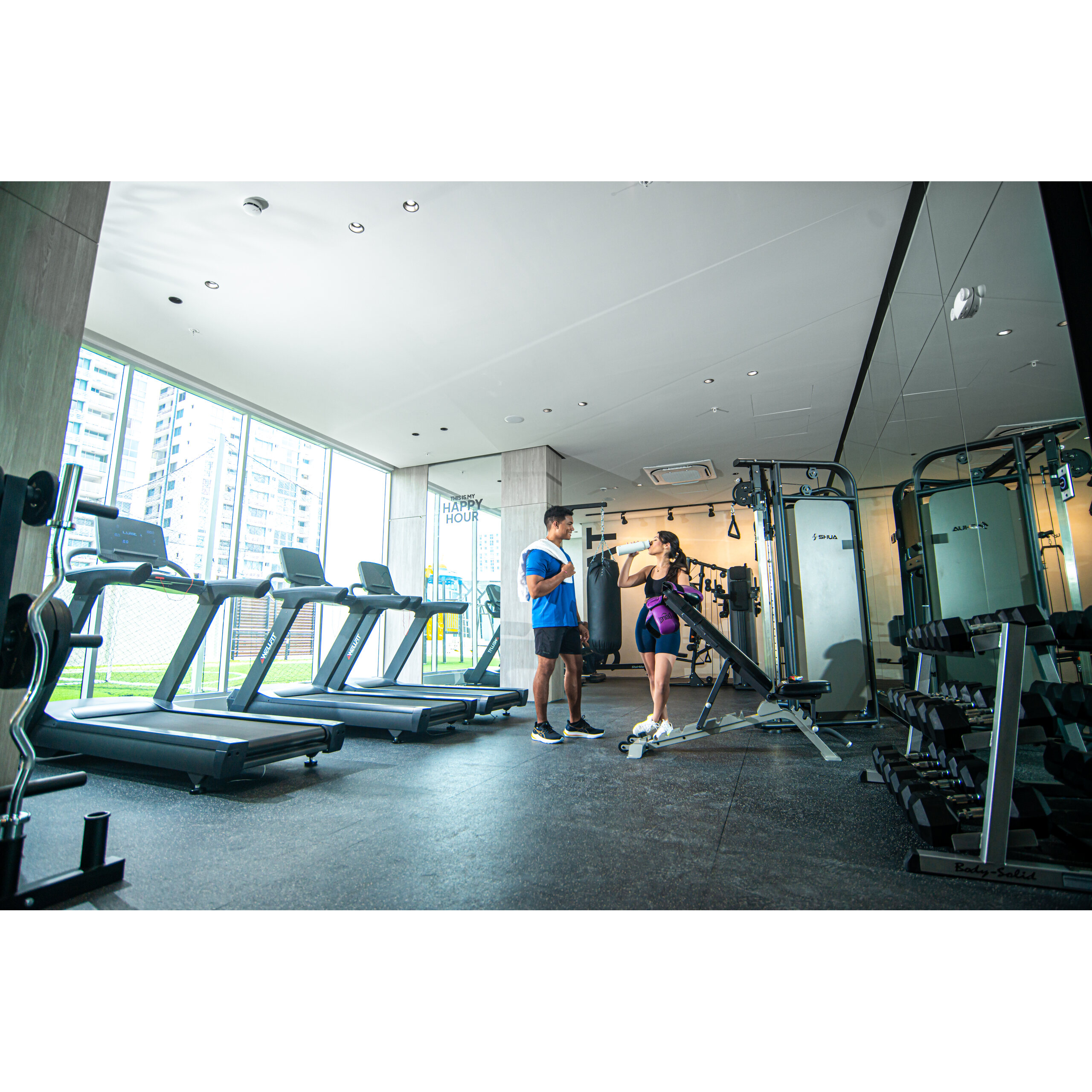 Gimnasio interior con caminadoras, pesas y dos personas conversando.
